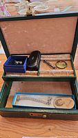 Vintage green jewelry box open showing assorted jewelry inside including earrings in blue box, bracelet in lower compartment, and decorative pins.
