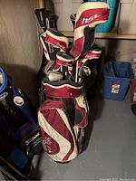 Golf bag in beige and maroon with multiple head covers inside and golf clubs, with a concrete wall background.