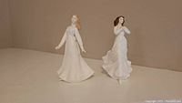 Two Royal Doulton figurines displayed side-by-side on a shelf with a plain background, showing their full figure and dresses