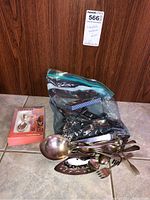 Plastic bag containing multiple silver-plated flatware pieces and a boxed item on left side on a tiled floor against wooden wall