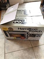 Hamilton Beach toaster oven box sitting on tile floor, showing front brand and product information.