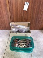 Mixed utensils in a green plastic container and a cardboard box, placed on tile floor with wooden background.