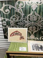 Photo showing a framed print of a stylized fish in red and black, artwork signed, set against a patterned green and silver background.