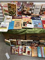 Photo showing a large collection of various used cookbooks arranged on a table and boxes below.
