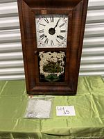 Full front view of the wooden framed clock on green cloth with painted glass lower panel and sealed bag of parts beside it.