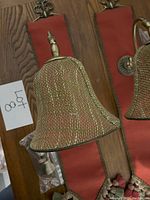 Pair of Italian wall sconces laid flat on wooden floor showing brass-tone mesh bell shades and red fabric panels with tassels and decorative finials. Plastic bags with plug-in cords visible.