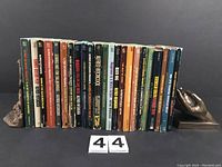 Front view of the 23 paperback books firmly stood upright with a holding hand bookend