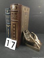 Front and spine view of both leather-bound books with a metal bookend shaped like hands, showing gold embossed titles and decorative details.