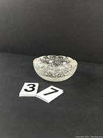 Front and angled side view of clear glass bubble ashtray with scalloped edges on black background.