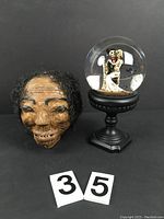 Front view of paper-mache shrunken head with detailed face and attached dark hair beside snow globe with calaca wedding couple inside on pedestal base, with lot number cards visible.