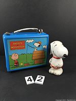 Blue metal lunchbox featuring Snoopy, Woodstock, and Charlie Brown, next to standing Snoopy moneybox figure.