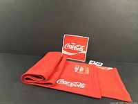 Folded red Coca-Cola scarf with white logos and graphics placed next to a matching large patch with the Coca-Cola logo embroidered.