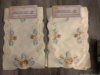 Two stacks of brand new cream-colored 12" x 18" embroidered placemats with scalloped edges and floral designs, still in packaging labeled Table Collection.