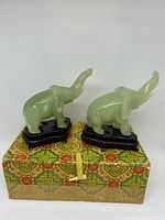 Two small polished jade elephant figurines with raised trunks displayed standing on black wooden stands on top of a decorative fabric-covered box with a printed multicolor pattern.