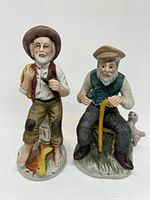 Front view of two vintage porcelain figurines, fisherman standing and elderly man sitting with dog.