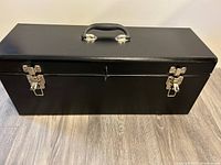 Front view of large black metal toolbox showing silver latch closures and padded handle on the top.