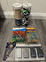 Photo showing two tall containers filled with mixed white, black, and grey beads, plus assorted color plastic beads in two 5 oz sealed packs at floor level.