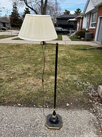 Full view of the floor lamp standing outside on a driveway with an off-white pleated shade showing its shape and crack inside.