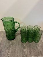 Full set of green glass pitcher and six matching glasses on a wooden floor against a white wall