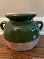Green two-handled pottery jar showing glossy green upper portion and natural bottom half, positioned on wooden floor.