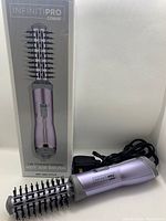 The Infiniti Pro Conair hot air brush outside of its box, showing the brush barrel with black bristles, light purple body and power cord coiled beside it.