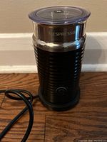 Front view of the black Nespresso Aeroccino milk frother showing the ribbed black plastic exterior, power button, and cord.
