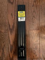 Full view of the black metal drywall roll lifter showing the rectangular base with grooves and holes, on a wooden floor.