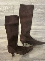 Side view of a pair of brown suede tall boots with kitten heels laid flat on wood flooring, one boot lying on its side showing heel and sole condition, the other standing upright.