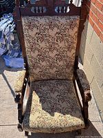 Front view of vintage wooden rocking chair showing floral upholstery on seat and back, with detailed wood carving on armrests and frame.