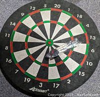 Top-down photo of round Accudart dartboard featuring black, white, red, and green segments. Set of 3 silver metal darts and 3 dart flights in clear plastic packaging placed on the board.