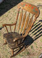 Brown wooden rocking chair outdoors on grass, showing floral design on backrest and condition of seat and armrests.