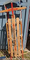 Full view of the American Eagle Fast Trak sled leaning against a light blue wall, showing wood slats and red metal runners and steering bar.