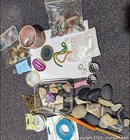 Overview of various stones and gemstones in containers, baggies, and spread out on paper. Includes red cup, plastic display box with small stones, and loose stones in different sizes and colors.