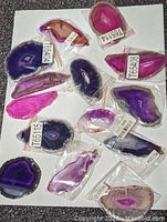 Top-down photo showing multiple polished agate slabs in varying hues of blue, purple, pink, and brown, each individually wrapped and labeled, arranged on a white background.