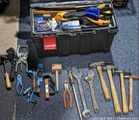 Photo showing a black plastic toolbox with one broken latch opened, various hand tools laid out around it including hammers, wrenches, pliers, a hand drill, C-clamp, and woodworking mallet.
