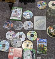 Photo showing Xbox game cases and discs spread on floor including Xbox, PS3, and Nintendo Switch games along with blank CDs and music CDs