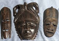 Three wooden African masks placed side by side on a textured fabric background showing detailed facial carvings and distinct styles.