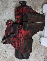 Photo of a heavy-duty metal vice painted red with signs of wear, showing the gripping jaws and metal handle. The vice includes a flat anvil surface on the back side.