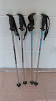 Four child size Rossignol ski poles standing upright against white wall, showing black rubber handles, attached wrist straps and gray shafts.