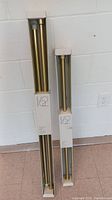 Two boxed gold curtain rods standing vertically against a white wall, showing the different sizes and packaging.