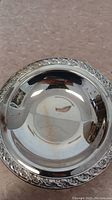 Small round silver shallow bowl with decorative floral border, polished and reflective surface.