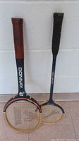 Two rackets hanging on wall, one tennis racket with Donnay branding and red grip, one badminton racket with blue frame and black grip.