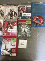 Photo showing 10 game cases arranged on a surface, featuring God of War III, SingStar titles, Batman, and NBA 2K14 discs out of case.