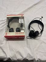 Photo showing one new in box and one open Puma The League on-ear headphones, both black with wired connection, lying on white cloth.