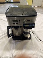Front and top view of a black and silver Flexbrew coffee maker with a glass carafe and single-serve cup. Dust visible on surfaces.