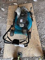 Top-down view of Honda lawnmower showing blue deck, engine, and handlebars resting on plywood board in gravel setting.