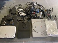 PlayStation consoles, controllers, and cables spread on a surface showing 2 PS1 consoles and 1 PS2 console with controllers and cables tangled