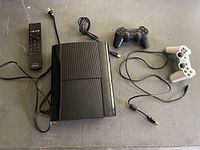 Sony PS3 Slim console with controllers, remote control, and cables arranged on floor showing top and side views.