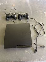 Top view of Sony PS3 console with two wired controllers and power cable laid out on floor.