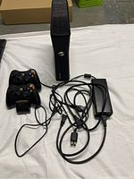 Full setup with Xbox 360 console standing vertically, two wired controllers on charging dock, power supply with attached cables all visible on white surface